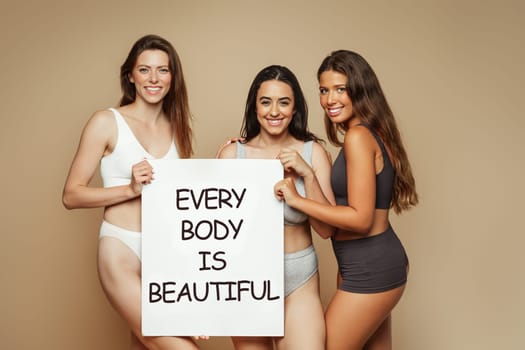 Smiling millennial diverse women in underwear enjoy lifestyle, hold banner with inscription every body is beautiful, isolated on beige background, studio. Motivation for self love, beauty care