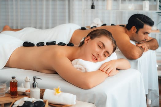 Hot stone massage at spa salon in luxury resort with day light serenity ambient, blissful couple customer enjoying spa basalt stone massage glide over body with soothing warmth. Quiescent