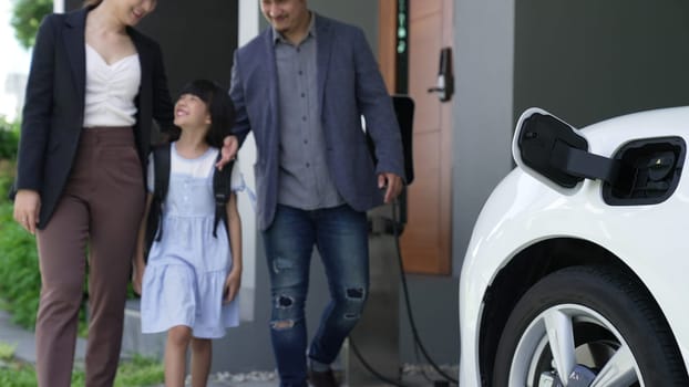 Progressive young parents and daughter with electric vehicle and home charging station. Green and clean energy from electric vehicles for healthy environment. Eco power from renewable source.