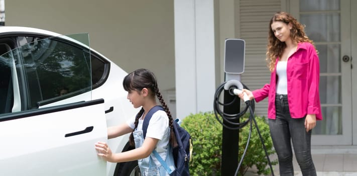 Happy little young girl learn about eco-friendly and energy sustainability as she help her mother recharge electric vehicle from home EV charging station. EV car and modern family. Panorama Synchronos