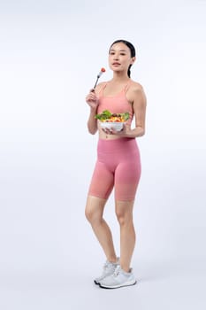 Young sporty Asian woman in sportswear holding salad bowl fill with vibrant of fruit and vegetable. Natural youthful and fit body lifestyle with balance nutrition on isolated background. Vigorous