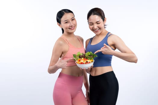 Two young sporty Asian women in sportswear holding salad bowl fill with fruit and vegetable. Natural youthful and fit body lifestyle people with balance nutrition on isolated background. Vigorous