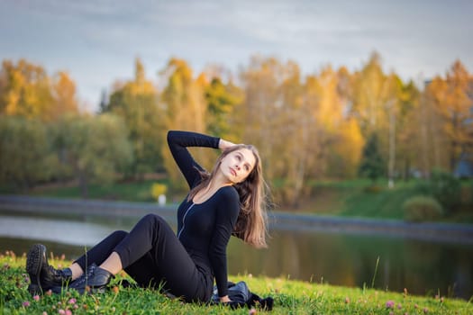 A beautiful girl poses while sitting on the grass by a pond in an autumn park