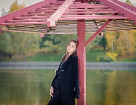 A beautiful girl poses while standing by a pond under an umbrella in an autumn park