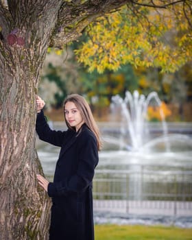 Portrait of a girl near a tree in a city park against the backdrop of fountains