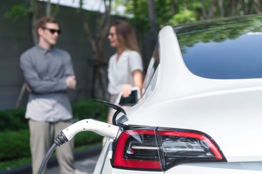 Young couple travel with EV electric car charging in green sustainable city outdoor garden in summer shows urban sustainability lifestyle by green clean rechargeable energy of electric vehicle innards