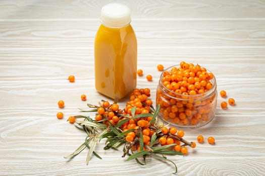 Sea buckthorn healthy drink in bottle, ripe berries in glass jar and branches with leaves top view on white wooden rustic background, great for skin, heart, vessels and immune system..