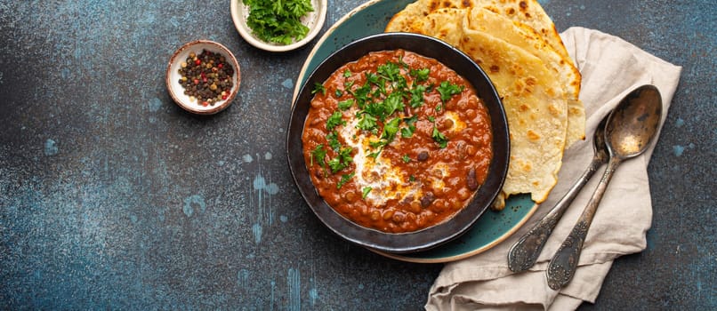 Traditional Indian Punjabi dish Dal makhani with lentils and beans in black bowl served with naan flat bread, fresh cilantro and two spoons on blue concrete rustic table top view. Space for text.