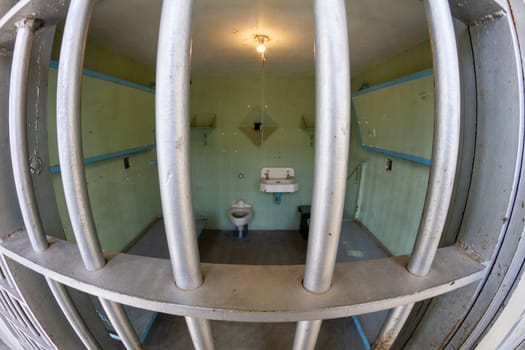 A jail cell with a toilet and sink. The cell is small and dirty. The bars on the window are rusty