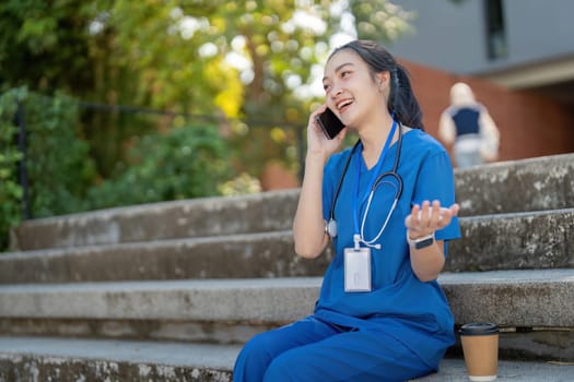 Asian nurse in blue scrubs having a lively phone conversation while sitting outside, displaying happiness.
