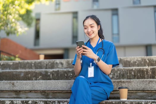 Cheerful Asian nurse in blue medical attire sitting on steps outdoors, using her phone during a break.