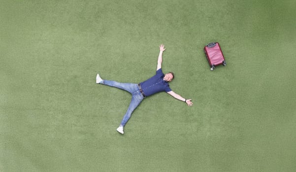 Happy man with a suitcase lies on a green lawn, top view. Arrival at the resort, vacation alone. Freedom from work