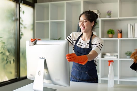 Asian woman wearing gloves cleaning a monitor in a home office, depicting a joyful mood and attention to detail.