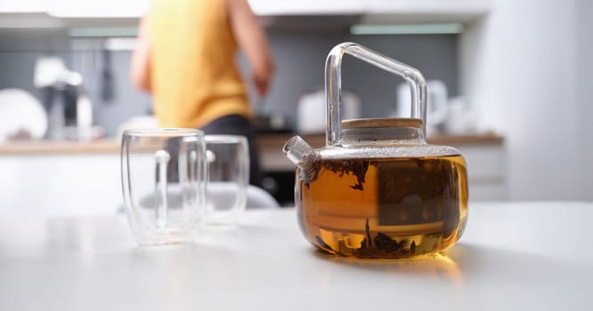 A woman made tea in a glass teapot, close-up, blurry. Glassware in the kitchen, coziness, hot herbal drink