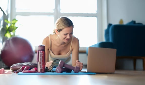 A woman makes a plank on her hands on the floor and looks into a laptop. Personal trainer, control, online training and motivation