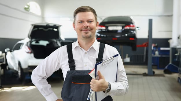 Portrait of qualified maintenance centre worker in special uniform posing on cars background. Mechanic male propose high service. Pit stop, garage concept