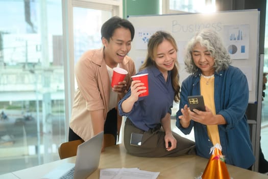 Cheerful corporate colleagues enjoying drink and looking at smartphone during an office party.
