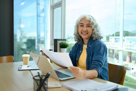 Confident senior businesswoman reviewing paperwork at her desk in modern office.