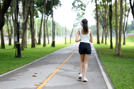 Back view of young woman wearing headphones jogging on pathway in the park.