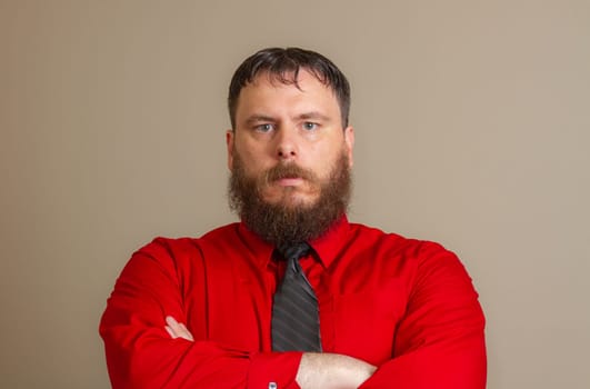 A man with a beard and a red shirt and a black tie is standing with his arms crossed