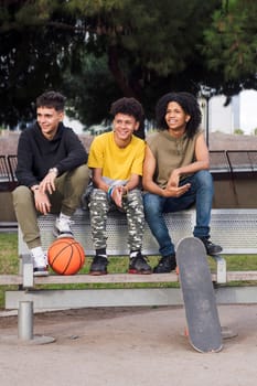 multiracial teen male friends smiling happy sitting on bench of a park with basketball and skateboard, concept of youth and urban lifestyle