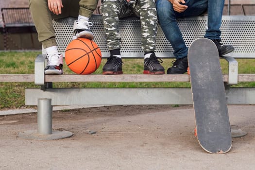 crop of three unrecognizable male friends sitting on bench of a park with basketball and skateboard, concept of youth and urban lifestyle