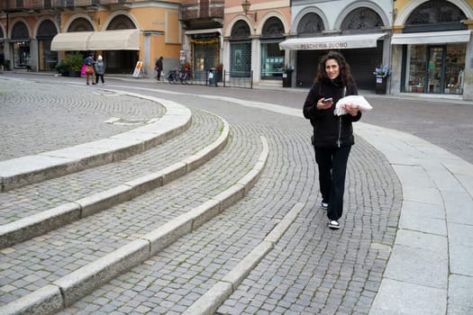 Cremona, Italy - November 20th 2024 Woman using smartphone and carrying takeout food descends cobblestone steps in piazza roma, cremona, italy