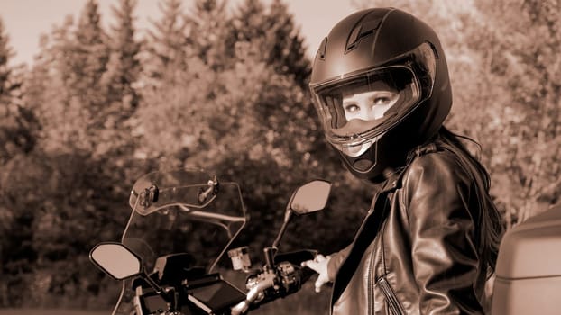 Young biker girl with touring motorbike on forest road. Colour of the year Mocha Mousse. Beautiful woman in leather jacket sitting on motorcycle