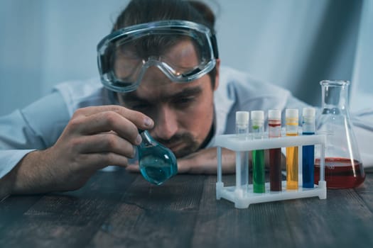 A scientist wearing protective goggles examines a round-bottom flask containing blue liquid, surrounded by test tubes and other lab equipment on a wooden surface. Chemistry research and analysis