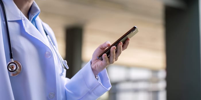 Asian doctor in a lab coat using a smartphone, representing communication and connectivity in a hospital setting.