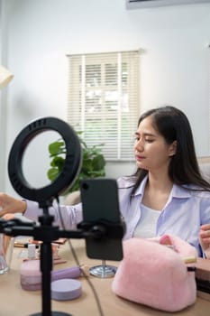 Asian woman beauty vlogger organizes makeup tools in front of camera, preparing content in a bright, home setting.