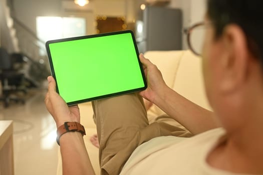 Over shoulder view senior man holding digital tablet with green screen sitting on the sofa at home.