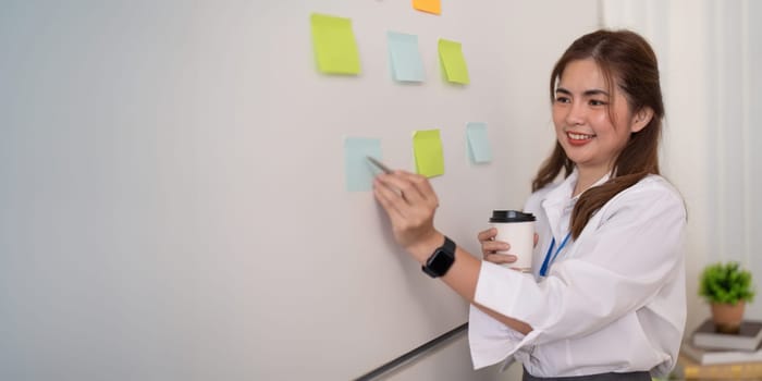 Businesswoman writing idea or tasks on sticky paper on wall, female team leader, executive manager, planning project, organize work.