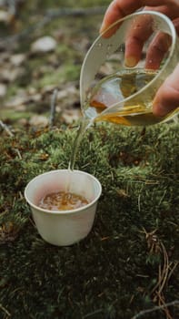 A hand pours golden liquid from a pitcher into a small cup in a peaceful outdoor setting surrounded by nature
