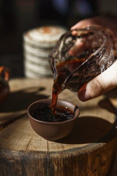 A skilled hand pours aromatic tea from a glass container into a rustic clay cup on a smooth wooden surface