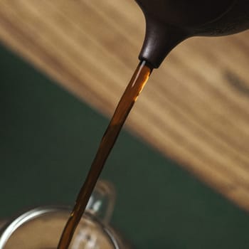 A closeup shot capturing the rich, dark brown coffee being elegantly poured from a kettle into a glass mug