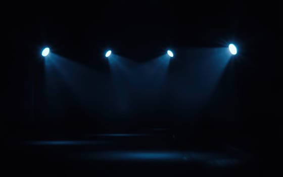 Three bright blue lights shine down on an empty stage, creating a dramatic atmosphere in the darkened venue, preparing for an upcoming performance.