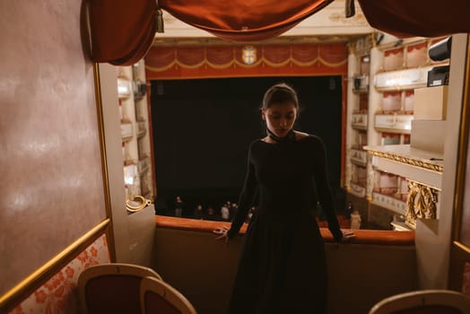 A woman in a stunning black dress stands gracefully at the theater balcony, observing the mesmerizing stage