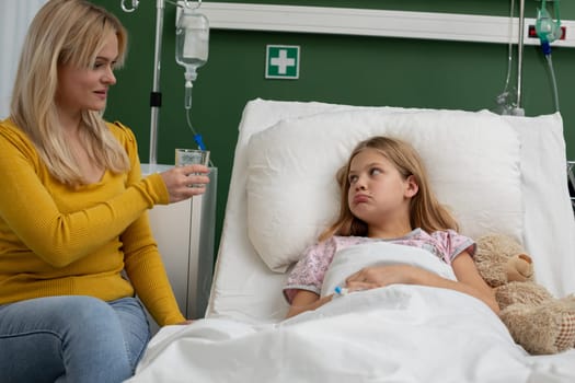 A caring and attentive mother gently offers water to her daughter, who is currently resting in a hospital bed