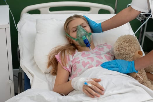 A young girl in a hospital bed uses an oxygen mask, comforted by a caregiver and her stuffed animal.