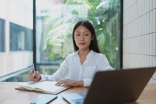 A young woman is seated at a wooden table in a bright and modern office, actively taking notes while using her laptop, representing focus and productivity in a contemporary workspace.