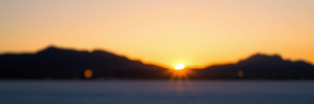 The sun rises slowly over mountain ranges, casting golden and orange hues across the calm lake's surface. Morning tranquility envelops the area as nature awakens.