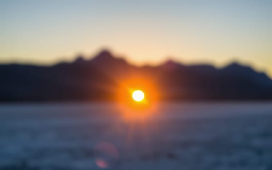 A vibrant sunset casts warm colors over rugged mountain silhouettes as the sun descends behind the horizon, creating a tranquil scene by the water.