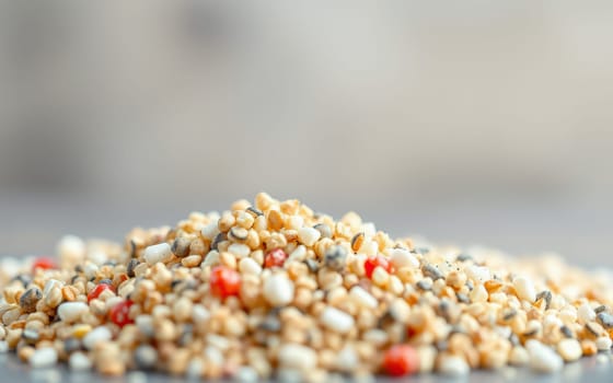 A close-up view shows a pile of assorted seeds on a flat surface, featuring tiny specks of red and hints of black throughout. The blurred background adds depth to the image.