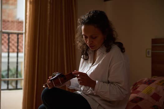 A woman in a white shirt tunes her guitar, seated near a window with gentle light illuminating the room. The atmosphere is peaceful and introspective, showcasing a moment of concentration.