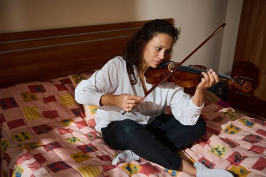 An adult woman in a casual setting playing violin, seated on a colorful bed. Natural lighting highlights her focus and the room's comfortable atmosphere.