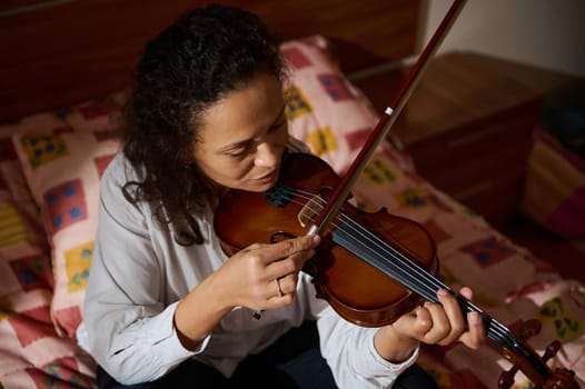 A focused woman plays the violin while sitting on a colorful bedspread in a warmly lit room, showcasing musical passion and home comfort.