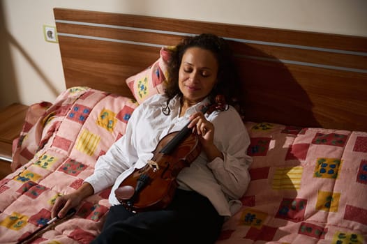 A woman enjoys a moment of relaxation in her bedroom, holding a violin. Her peaceful expression complements the warm, cozy atmosphere and colorful bedding.