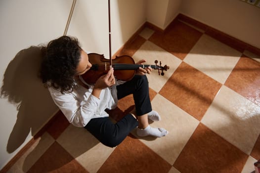 A musician seated on the floor plays the violin in a peaceful, sunlit room. The natural lighting enhances the tranquil atmosphere, creating a sense of calm and introspection.