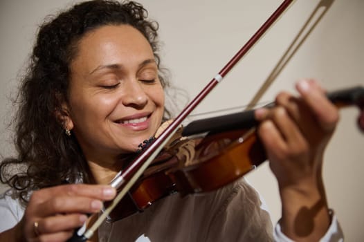 A woman is happily playing the violin, eyes closed and smiling, feeling the music and expressing joy.